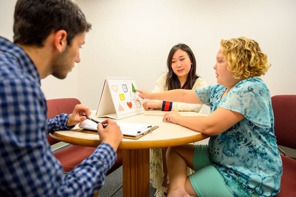 Two psychology students sitting with a young girl and taking notes while she points out items on a card in front of her