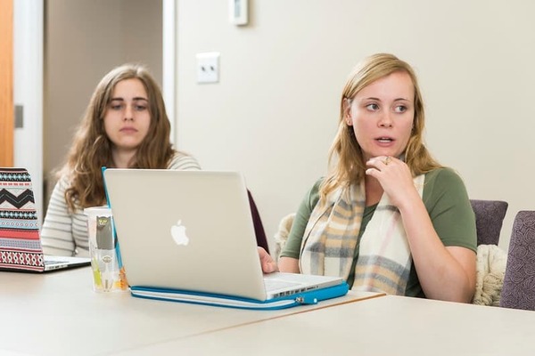 Graduate student sitting at a table behind a laptop looking and speaking to someone to the right