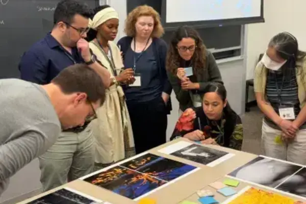 Diverse group intently examines art prints and sticky notes laid out on a table in a classroom setting, appearing engaged.